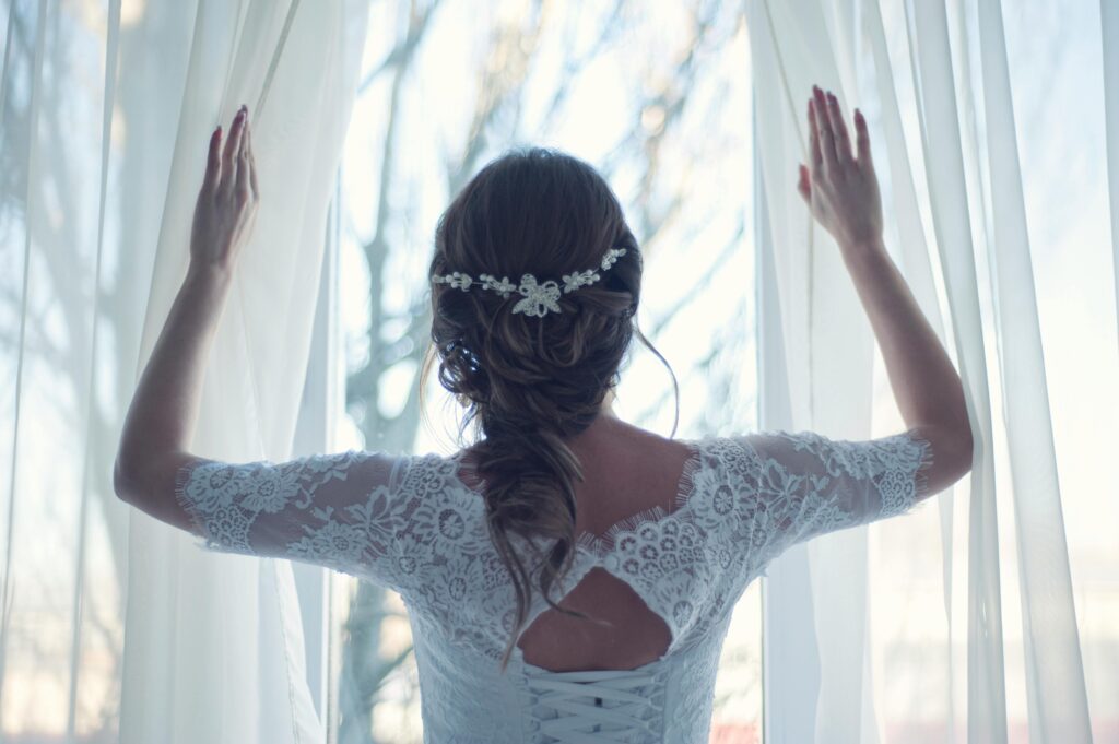 pexels-photo-341372-341372 Beautiful bride in lace dress looks out a sunlit window, capturing a moment of serene elegance.